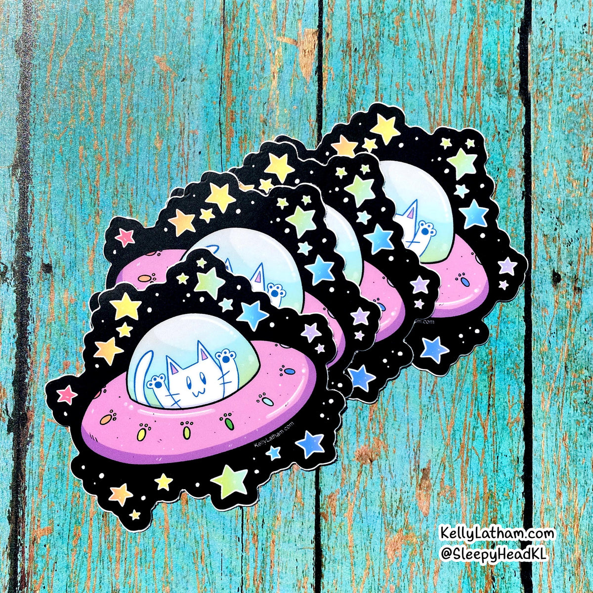 Three die-cut stickers featuring a cute cartoon white cat inside a pink UFO surrounded by stars, arranged on a distressed turquoise wooden background. The stickers have black scalloped borders and are signed by Kelly Latham and @SleepyHeadKl.