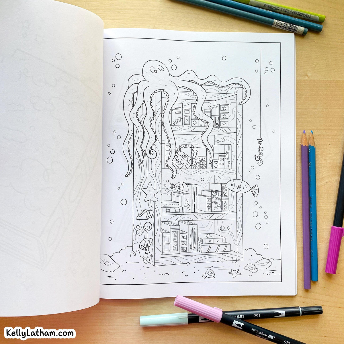 An open coloring book or art journal on a wooden desk, showing a black-and-white underwater scene page with an octopus, fish, bubbles, and a house. Colored pencils and markers are placed around it, and the watermark KellyLatham.