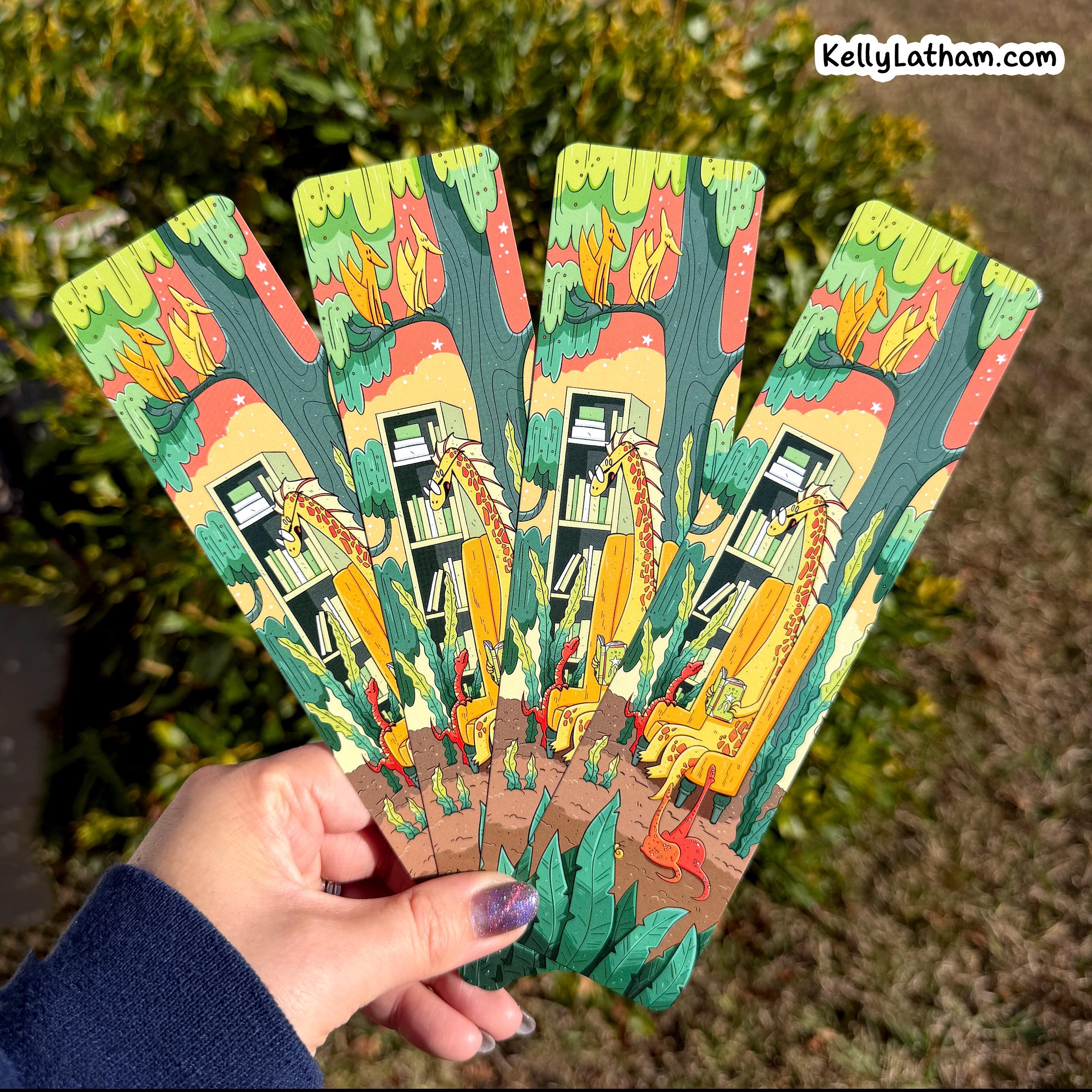 A hand holds a fanned-out set of four decorative bookmarks outdoors, with green foliage in the background. Each bookmark features a colorful illustration of a cozy library scene with books, shelves, and whimsical characters.