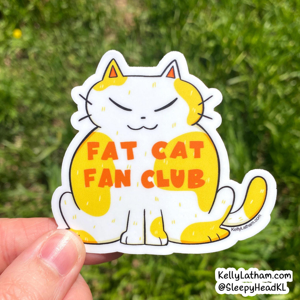 A hand holds a die-cut sticker of a cartoon cat with yellow patches and orange text reading 'FAT CAT FAN CLUB' against a blurred green grass background.