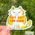 A hand holds a die-cut sticker of a cartoon cat with yellow patches and orange text reading 'FAT CAT FAN CLUB' against a blurred green grass background.