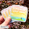 A hand holds a fan of square stickers outdoors, with blurred autumn foliage in the background. The front sticker reads “REMEMBER SELF CARE.