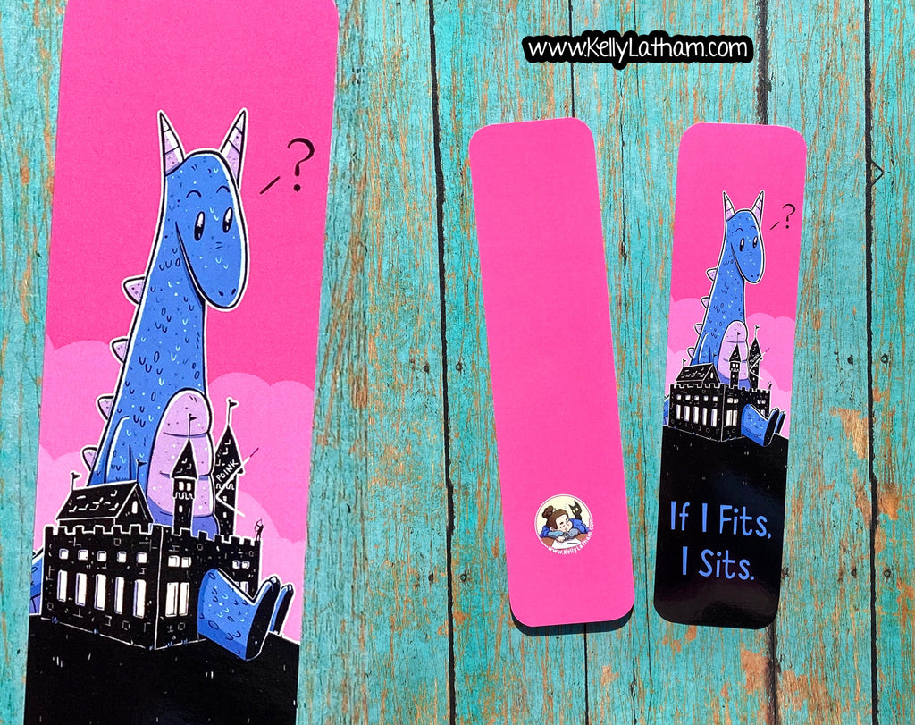 Three pink paper bookmarks with whimsical illustrations of a blue dragon and castle are arranged on a distressed turquoise wooden surface. One bookmark is shown flat with the full design, two are folded, and one displays the text 'If I Fits.