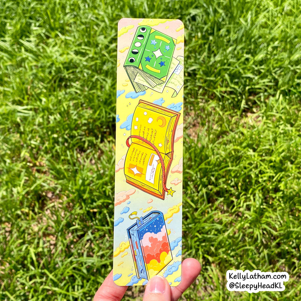 A hand holds a long, narrow decorative card or bookmark featuring three colorful illustrated books against a bright yellow background, with green grass blurred in the background. A small watermark in the bottom right reads KellyLatham.