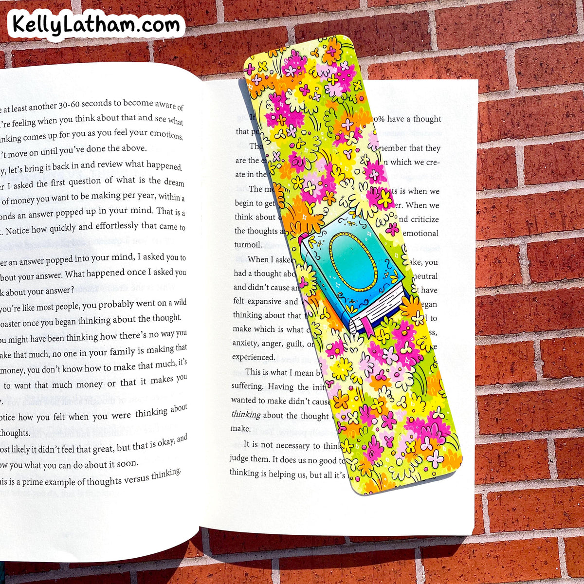 A colorful floral bookmark with a turquoise heart-shaped center and gold glittery text reading 'KELLY LATHAM' lies across open book pages on a red brick background.