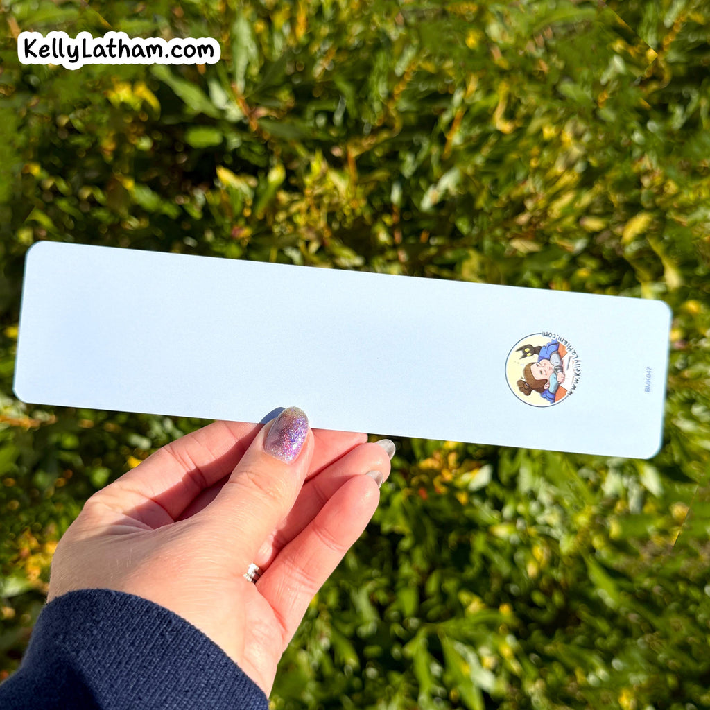 A hand with purple glitter nail polish holds a long, narrow light blue card or label outdoors against a background of green foliage. The card has a small circular cartoon logo on the right side and a white 'KellyLatham.