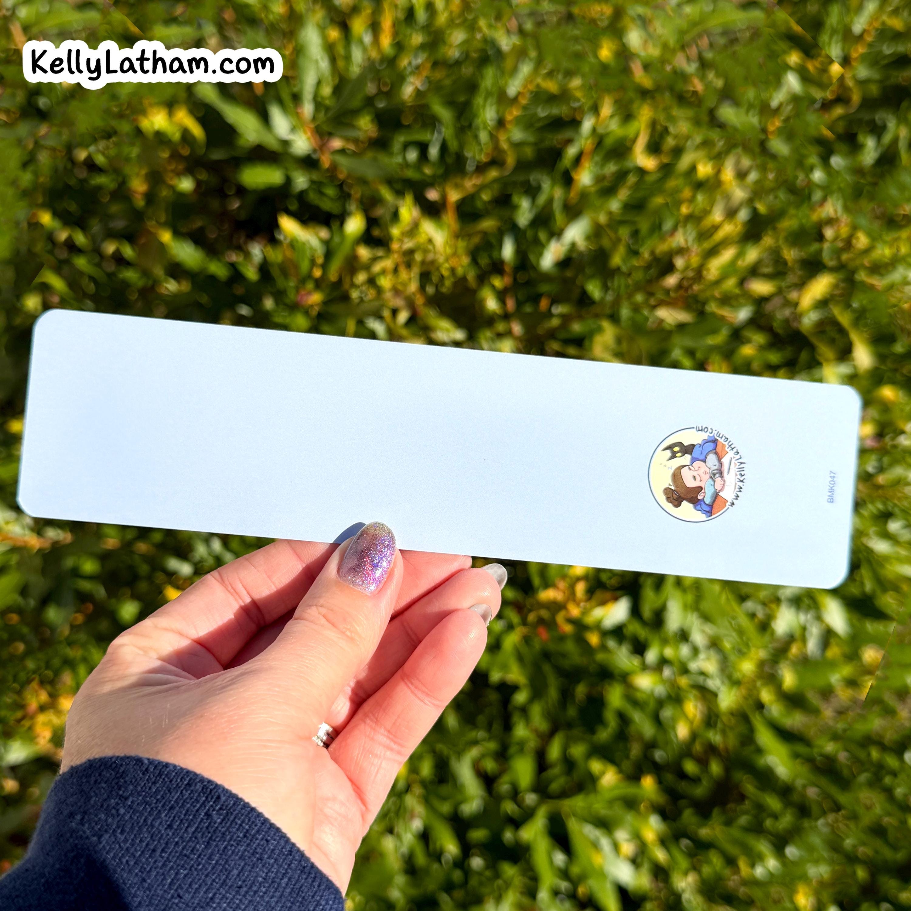 A hand with purple glitter nail polish holds a long, narrow light blue card or label outdoors against a background of green foliage. The card has a small circular cartoon logo on the right side and a white 'KellyLatham.