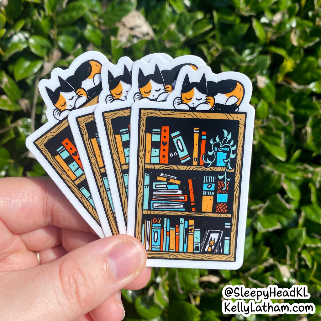 A hand holds a fanned-out set of die-cut stickers featuring a cartoon cat sleeping above a bookshelf with colorful books and plants. The background is blurred green foliage.