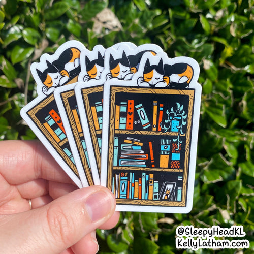 A hand holds a fanned-out set of die-cut stickers featuring a cartoon cat sleeping above a bookshelf with colorful books and plants. The background is blurred green foliage.