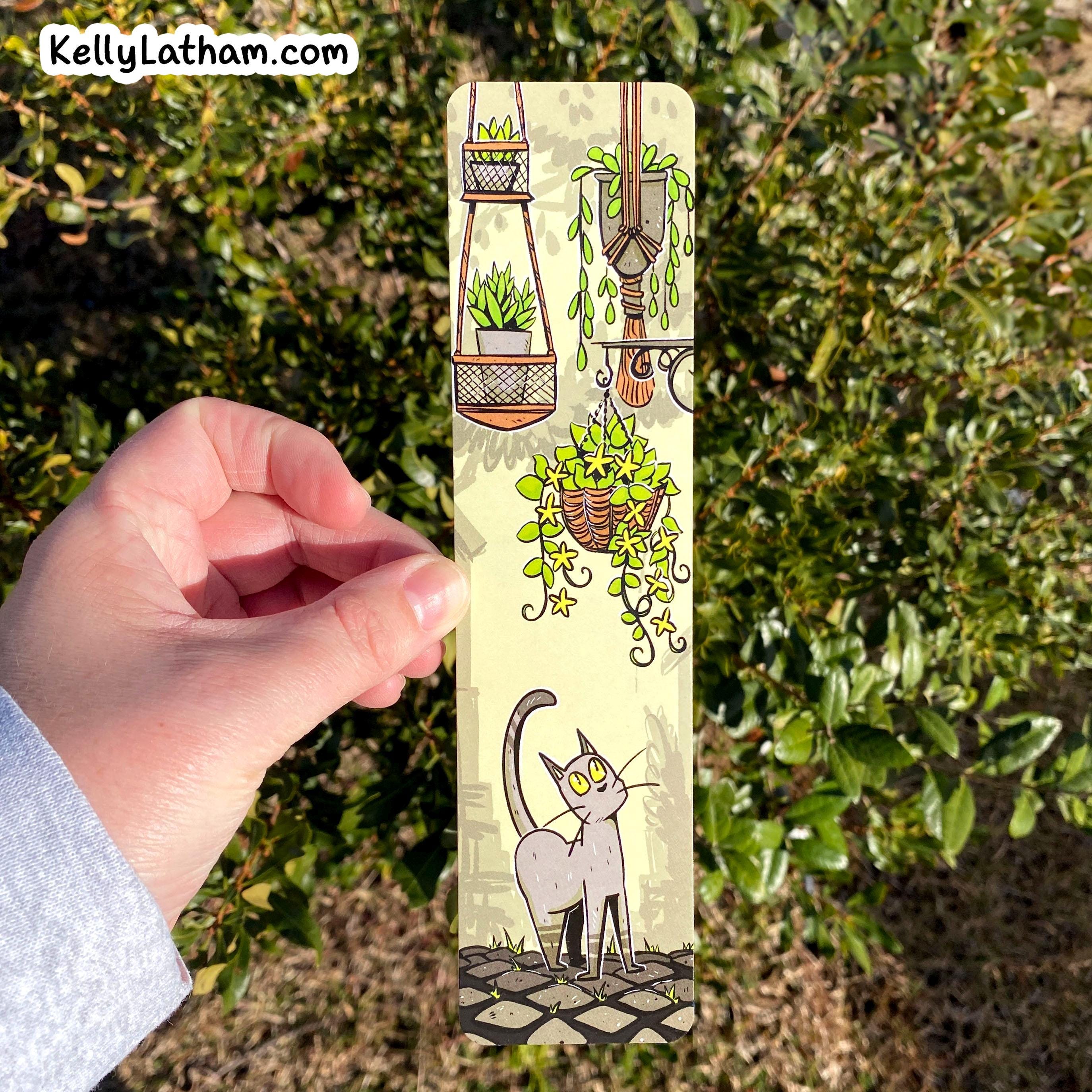 A hand holds a slim rectangular bookmark featuring a whimsical illustration of hanging plants and a gray cat on a cream background, photographed outdoors in front of green foliage.