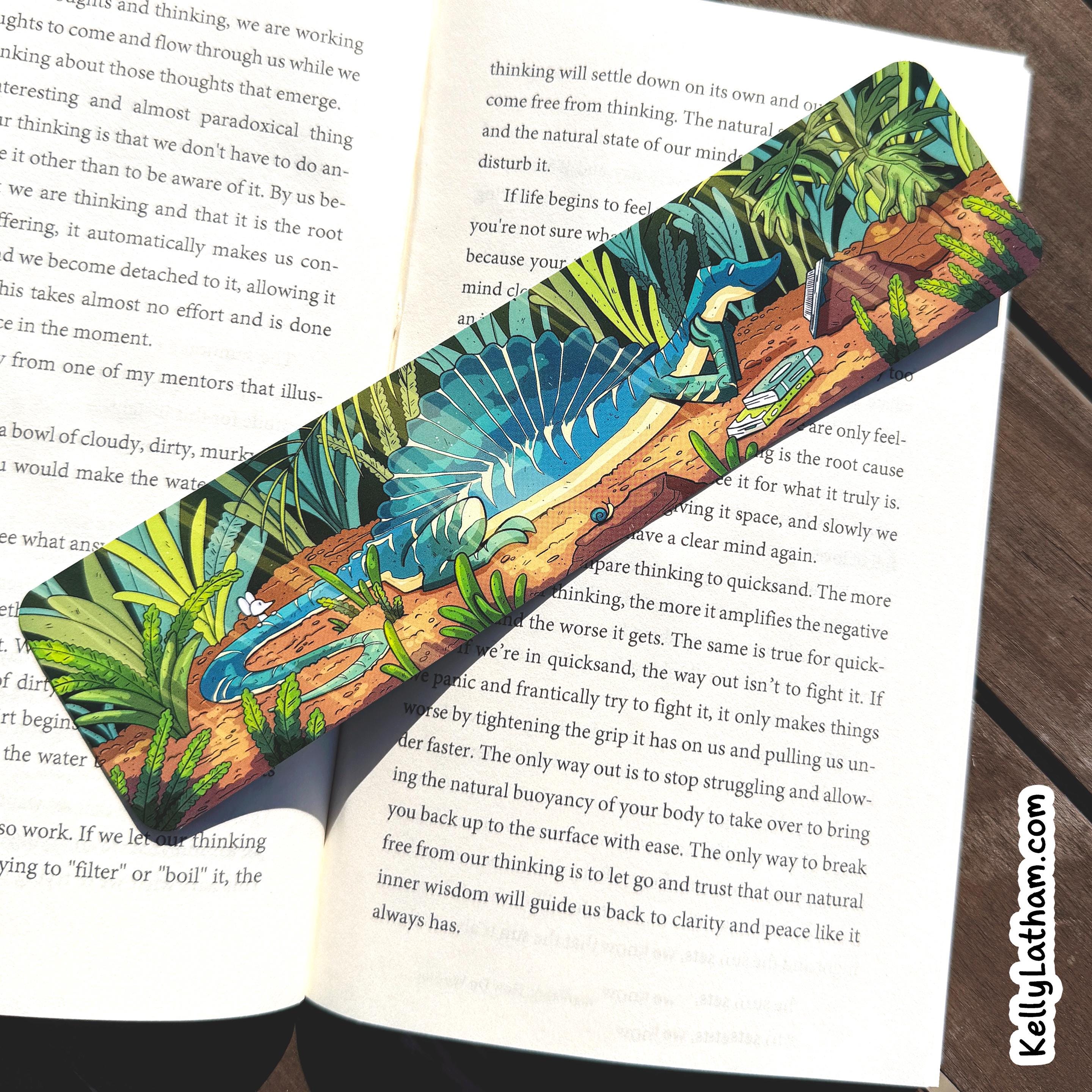 A colorful tropical leaf patterned bookmark or ribbon laid diagonally across an open book on a sunlit surface. The item has bright green, blue, orange, and yellow leaves with a turquoise base.