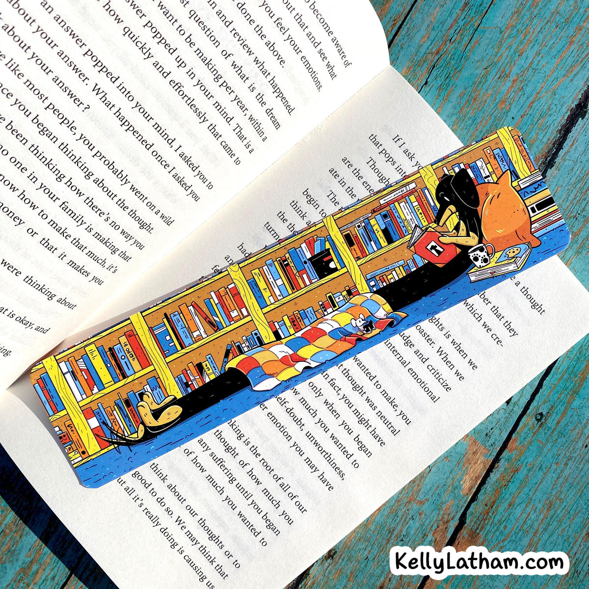 A 3D pop-up book bookmark shaped like a library scene with colorful bookshelves, books, a librarian, and a character, displayed on an open book over a distressed teal wooden surface.