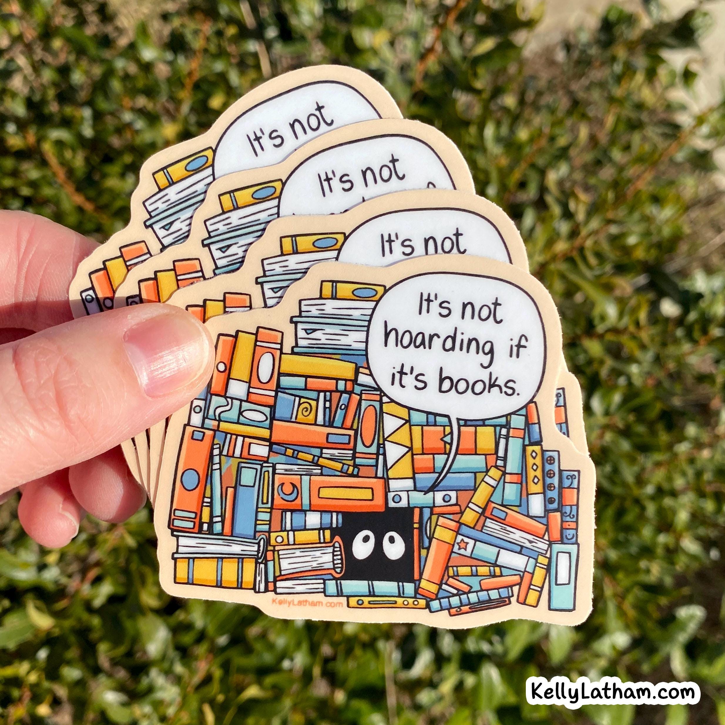 A hand holds a stack of die-cut wooden stickers outdoors against a blurred green hedge background. The stickers feature an illustrated pile of colorful books with a small black face and a speech bubble that reads, "It's not hoarding if it's books.