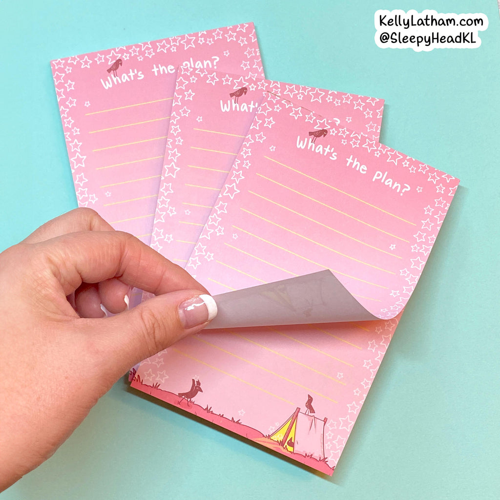 Three pink notepad sheets with a cute camping theme, featuring a hand peeling back the bottom cover to reveal a gray backing. The notepads have white text reading 'What's the plan?' and are decorated with stars, campfire, and tents.