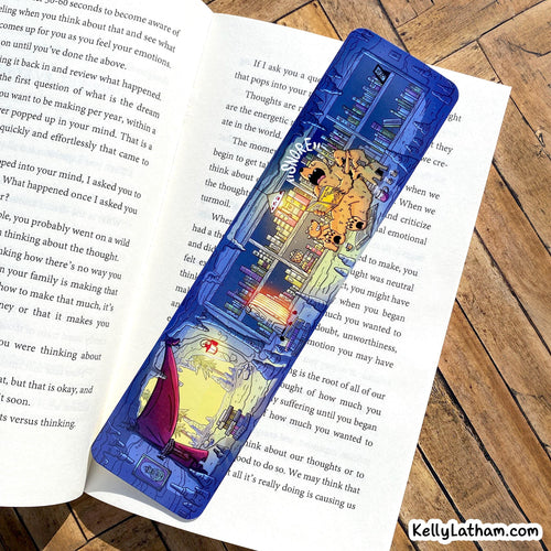 A blue and white illustrated pencil case featuring a cartoon character and the words 'Be Kind, Stay Kind,' placed on top of an open book on a wooden surface.