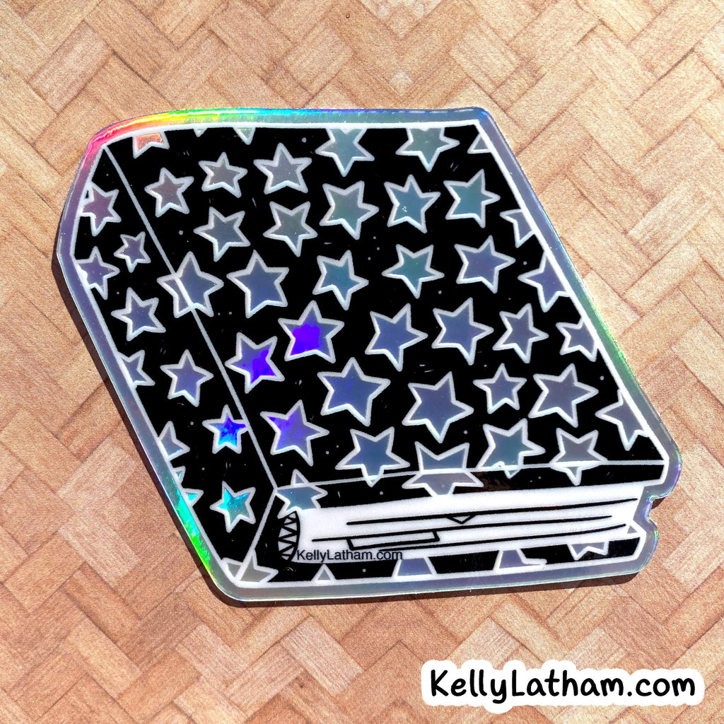 A die-cut sticker featuring a black background covered in silver and blue-gray stars, with a white horizontal line across the bottom and a small black scribble on the left. The sticker has a holographic iridescent border and is photographed on a light wood herringbone surface.