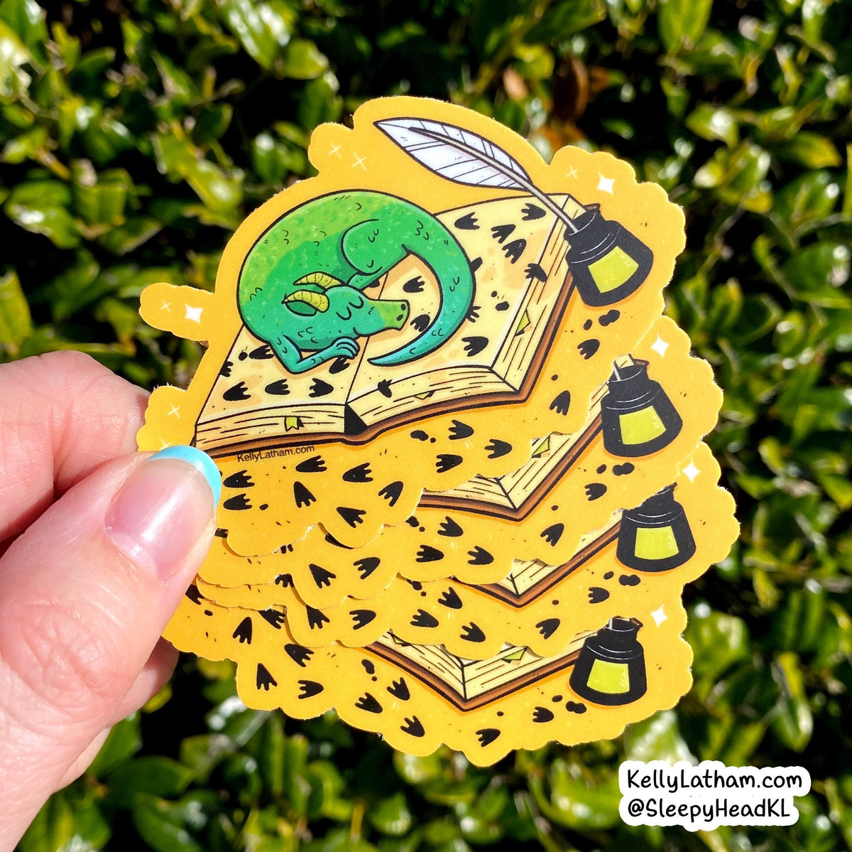 A hand holds a stack of yellow die-cut stickers featuring a green dragon curled up inside an open book, with quills and lanterns, against a blurred green hedge background.