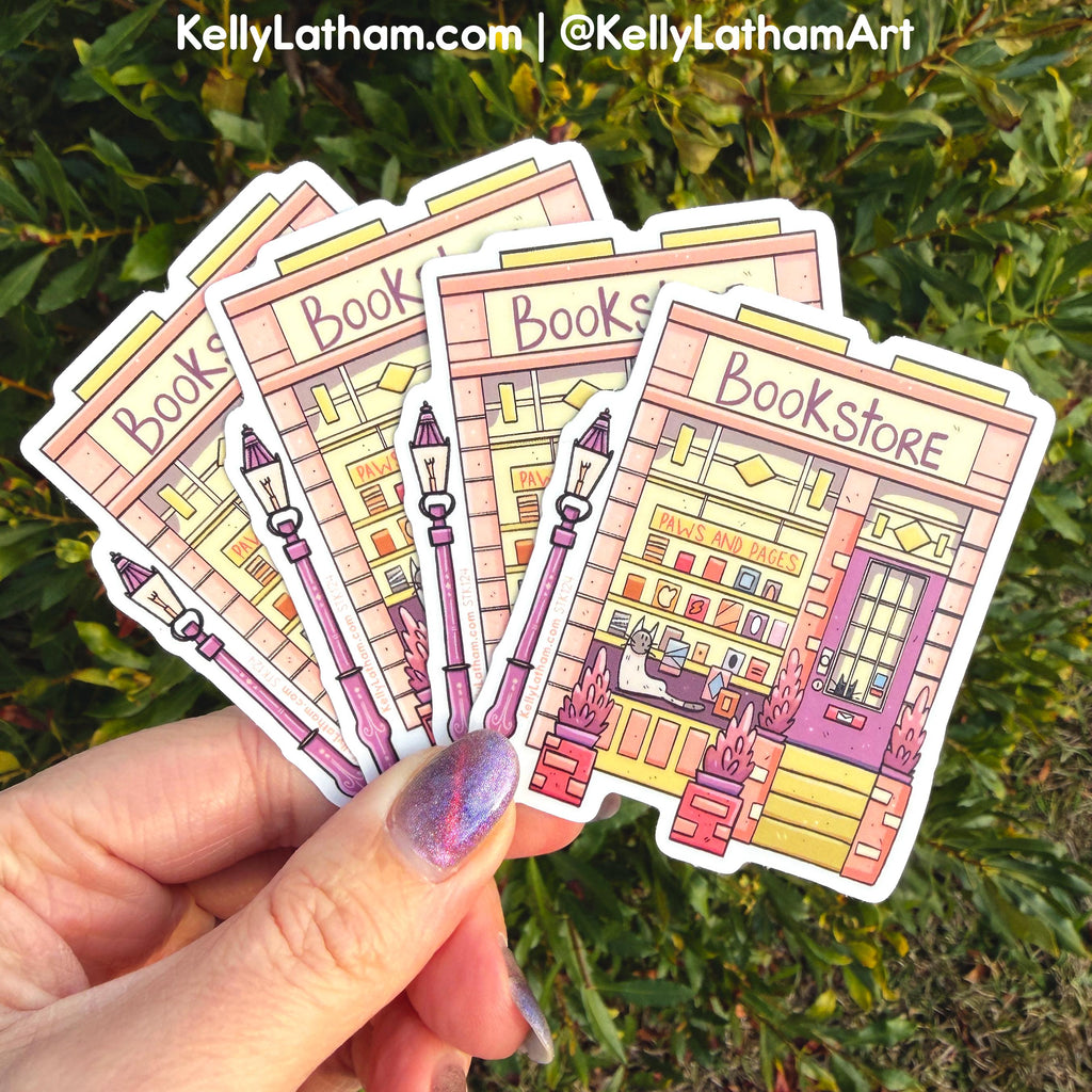 A hand with purple glitter nail polish holds five die-cut vinyl stickers depicting a colorful illustrated bookstore exterior, set against a background of green outdoor foliage. A watermark at the top reads 'KellyLatham.