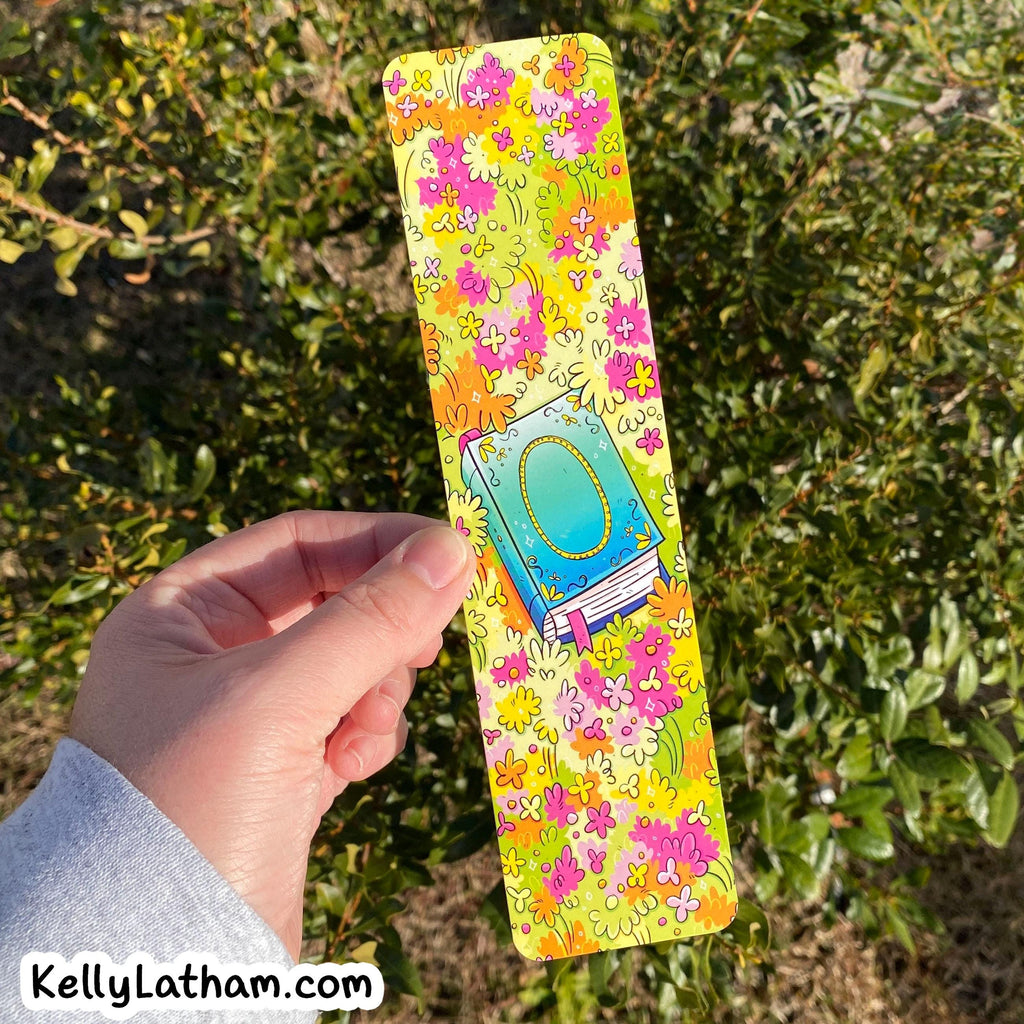 A hand holding a long rectangular card or bookmark featuring a colorful floral pattern and a central illustration of a blue book with a gold oval frame, set against a background of green outdoor foliage.