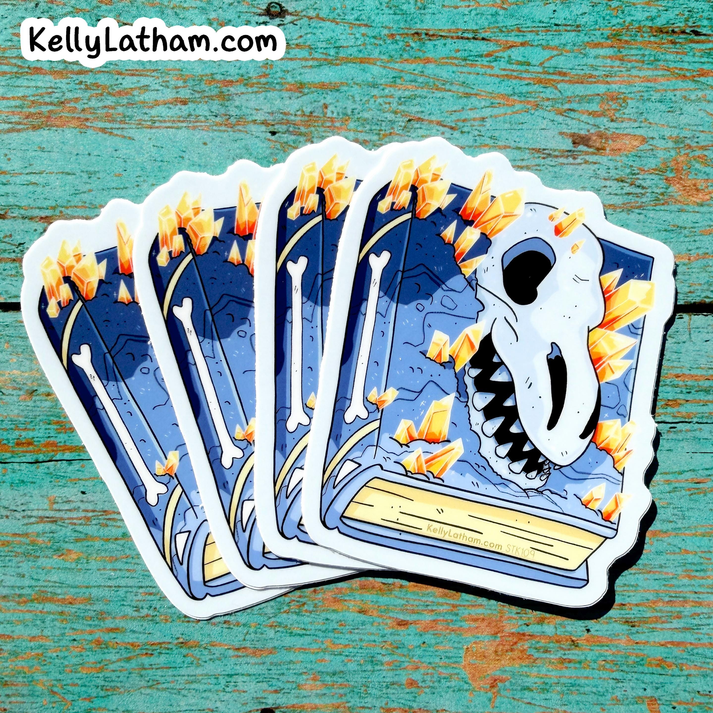 Four die-cut stickers featuring a blue and white illustrated design with a skull, bones, and orange crystals, fanned out on a distressed teal wooden surface. The brand name KellyLatham.
