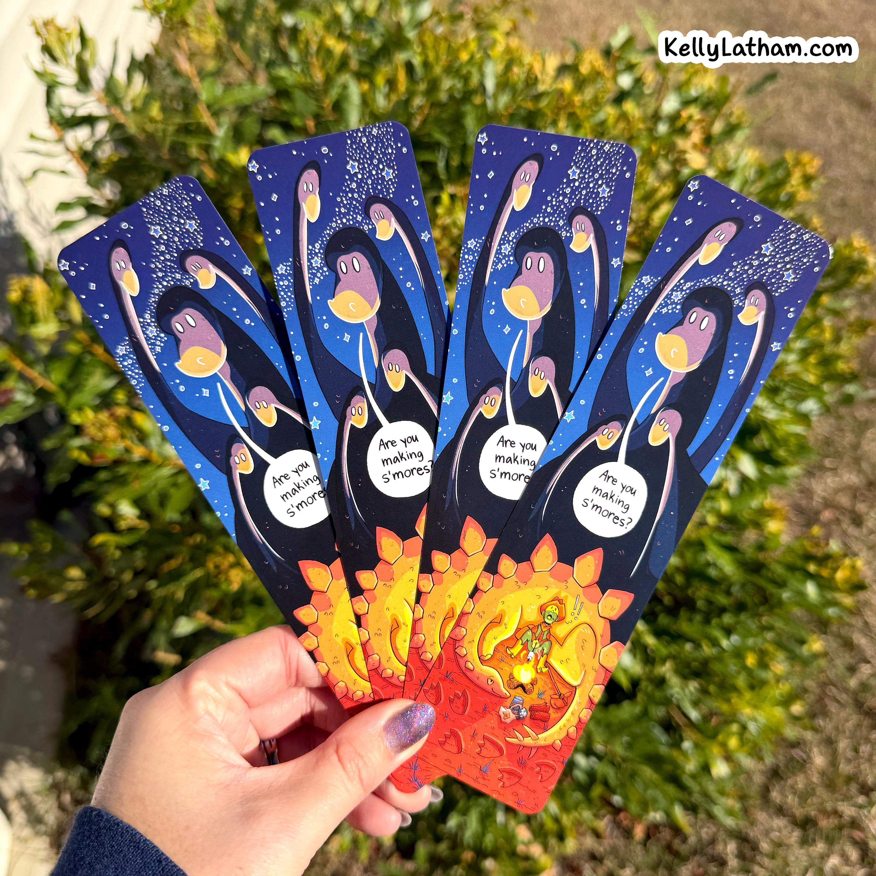 A hand holds four illustrated bookmarks outdoors in front of green bushes. The bookmarks are dark blue with cartoon penguins, white speech bubbles, and a red-to-yellow gradient at the bottom.