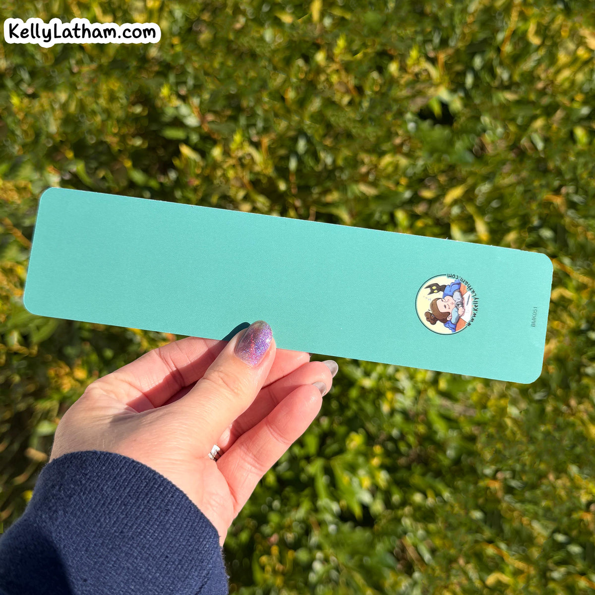 A hand holding a long, rectangular turquoise card or bookmark outdoors against a blurred green hedge background. The card has rounded corners, a small circular cartoon character sticker on the right side, and a visible 'KellyLatham.