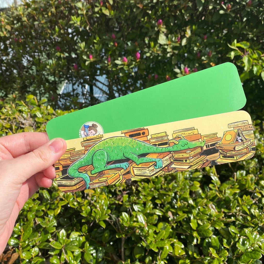 A hand holds a green rectangular card or sticker outdoors in front of dense green foliage. The lower portion of the card shows an illustration of a green chameleon resting on a pile of books, with a small circular sticker visible on the left side.