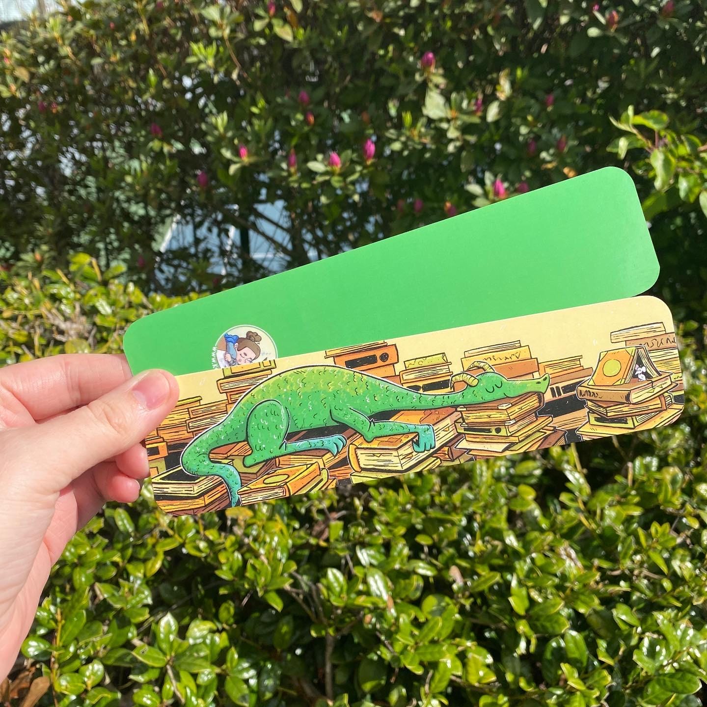 A hand holds a green rectangular card or sticker outdoors in front of dense green foliage. The lower portion of the card shows an illustration of a green chameleon resting on a pile of books, with a small circular sticker visible on the left side.