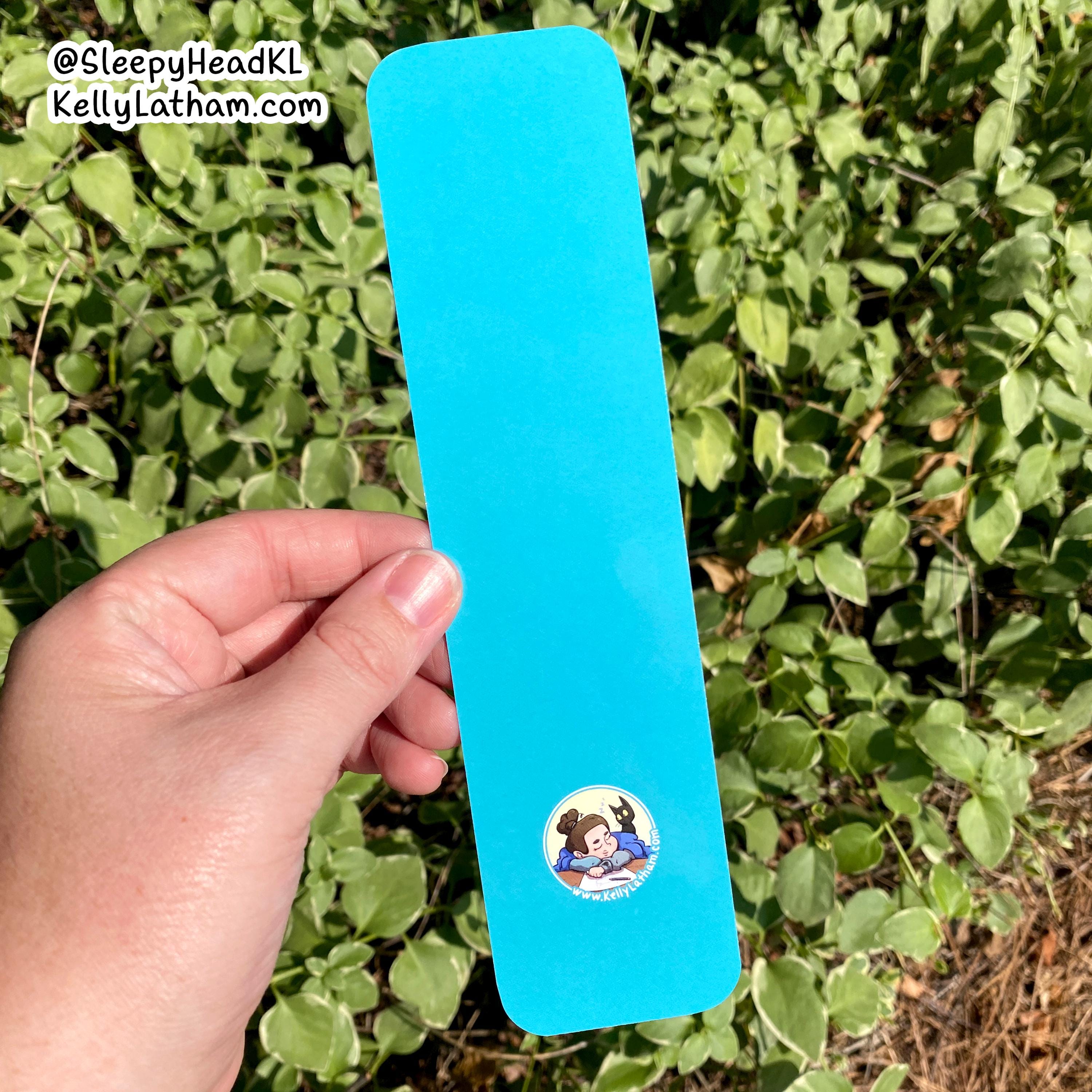 A hand holds a bright blue rectangular sticker with rounded corners against a background of green leaves. The sticker features a cartoon illustration of a brown-haired character in a blue swimsuit and a circular 'Peanuts' logo at the bottom.
