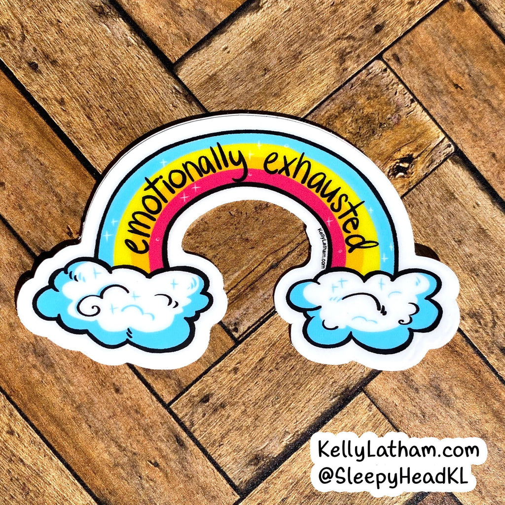 A die-cut sticker featuring a pastel rainbow with the words 'emotionally exhausted' in black script, resting on a brown wooden surface. The sticker includes two white clouds with blue shading at the bottom.