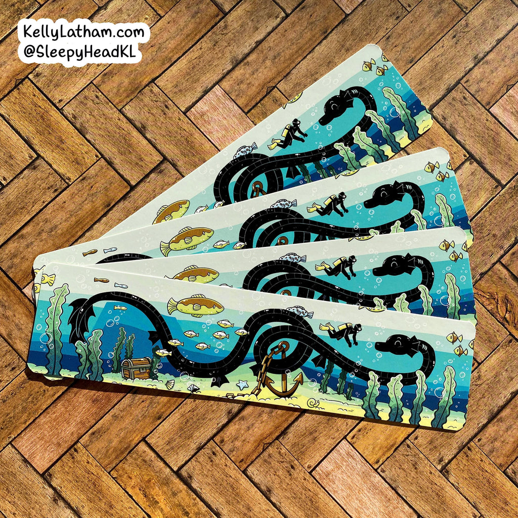Four rectangular coasters with an underwater scene print, fanned out on a brown wooden parquet floor. The coasters feature a blue ocean, black spiral track, yellow fish, and sea plants.