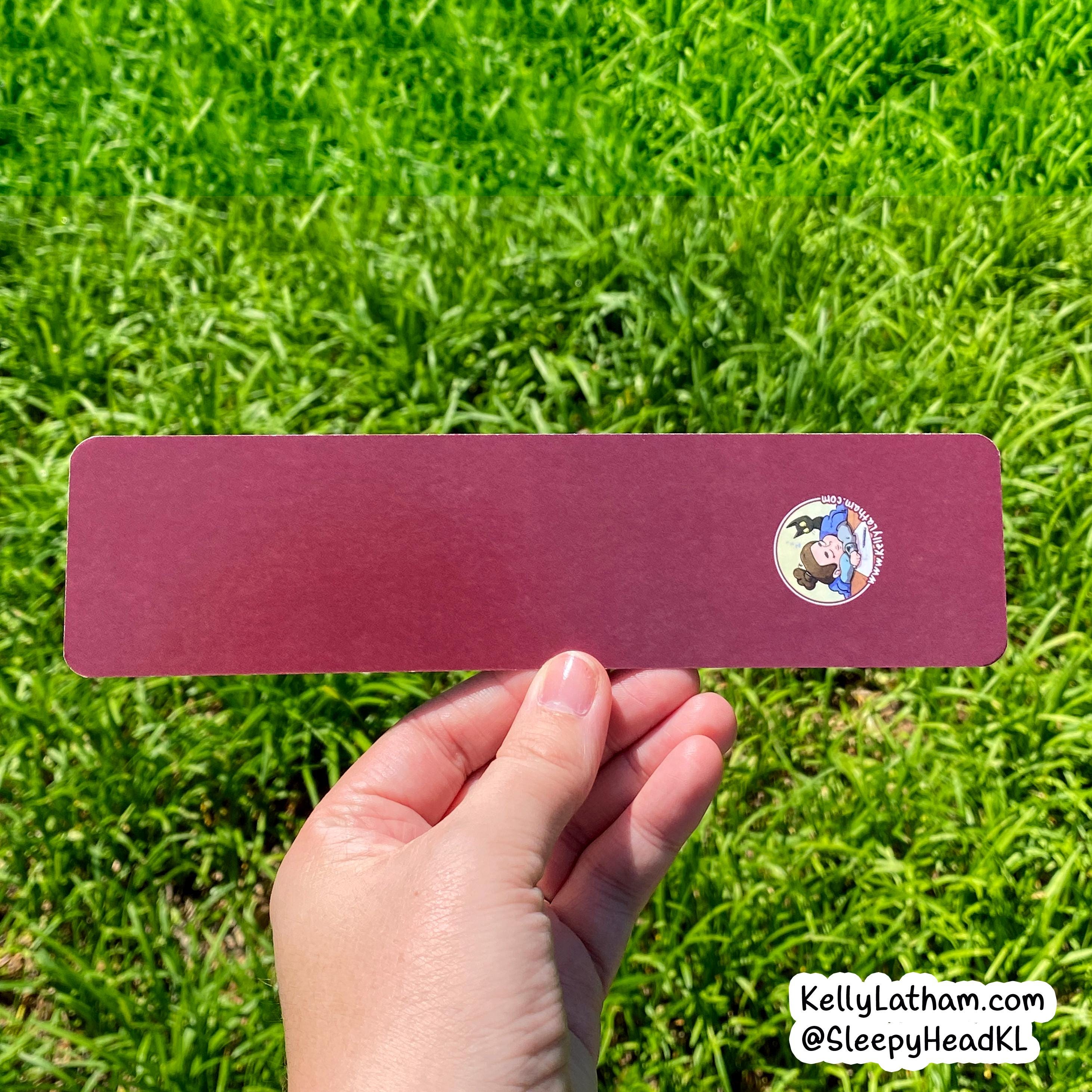 A hand holds a rectangular burgundy card or sticker against a background of green grass. The card has a small circular cartoon sticker on the right side and a white watermark in the bottom right corner.