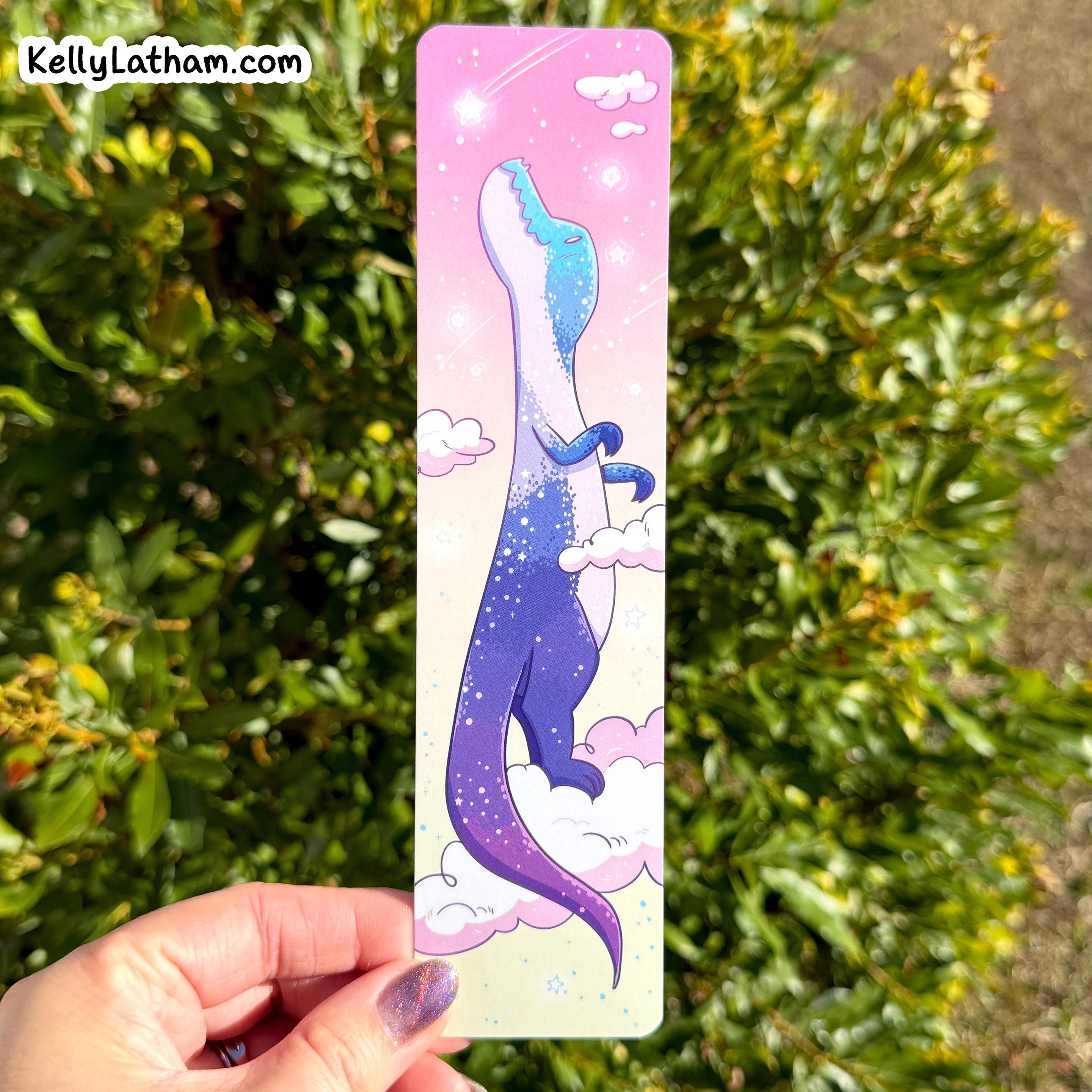 Bookmark with a colorful dinosaur design held by a hand against a green leafy background