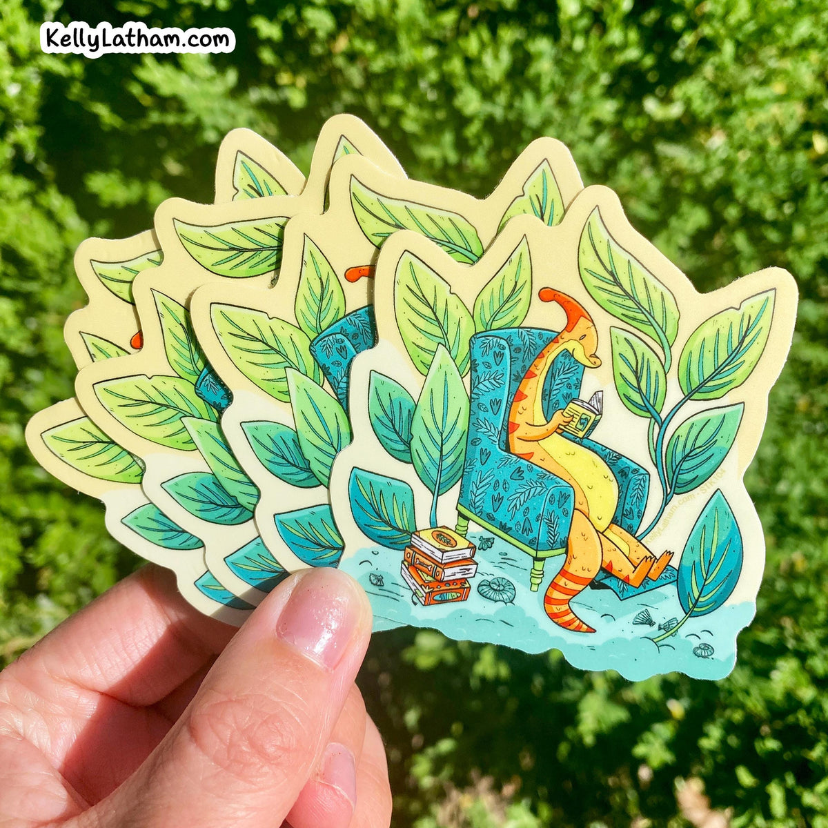 A hand holds a fanned-out set of die-cut stickers featuring a whimsical illustration of a yellow dinosaur sitting in a blue armchair surrounded by green leaves, with a blurred green outdoor background.