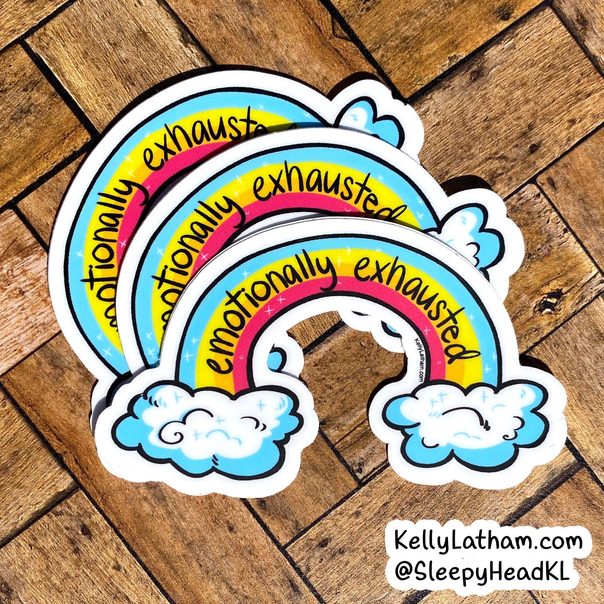 Three die-cut vinyl stickers of rainbows with the text 'emotionally exhausted' in black script, arranged on a brown wooden background. A watermark in the bottom right corner reads 'KellyLatham.