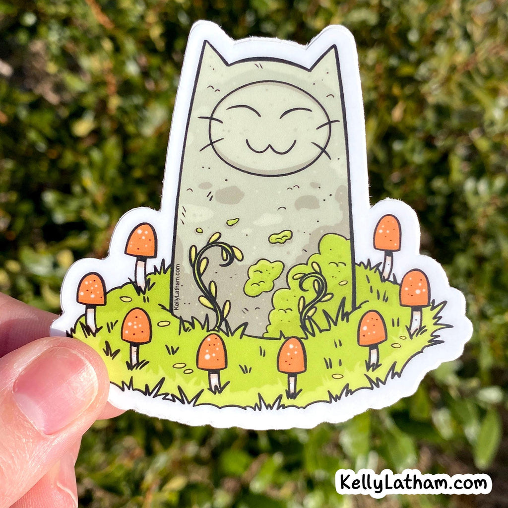 A hand holds a die-cut sticker featuring a cute cartoon cat standing in a grassy field with orange mushrooms, photographed outdoors against a blurred green background. The sticker has a visible KellyLatham.
