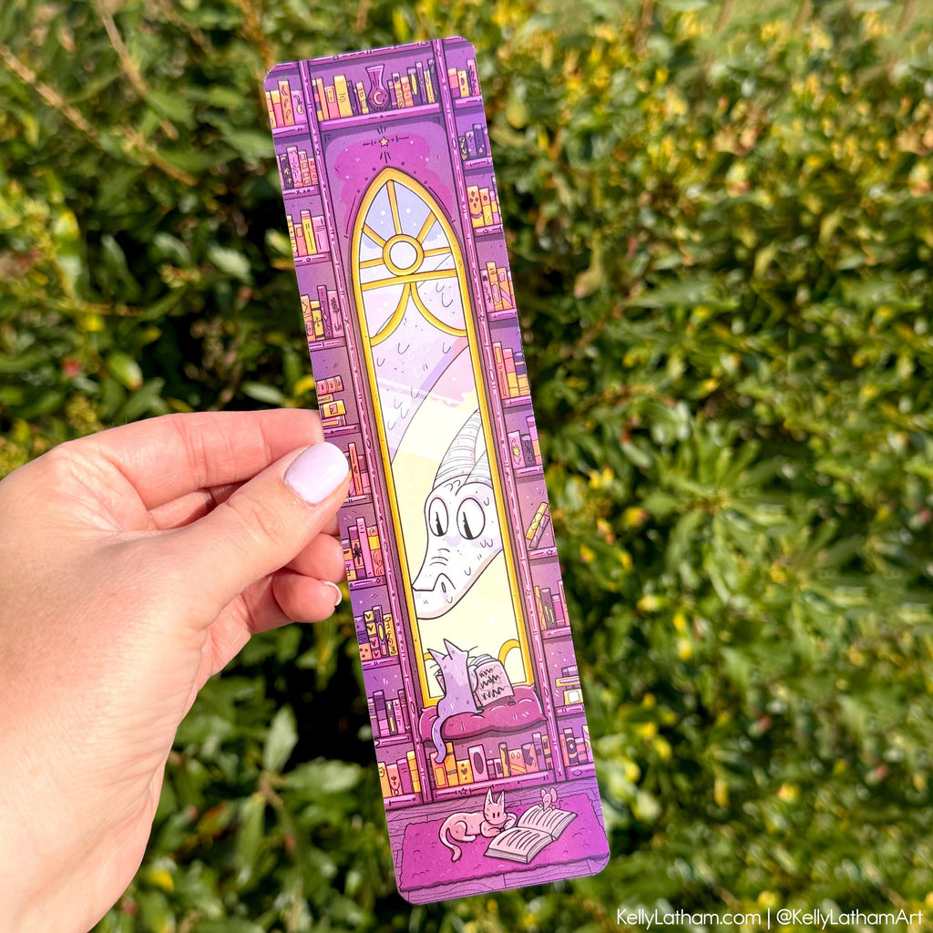 Hand holding a purple bookmark with cartoon design against a green leafy background