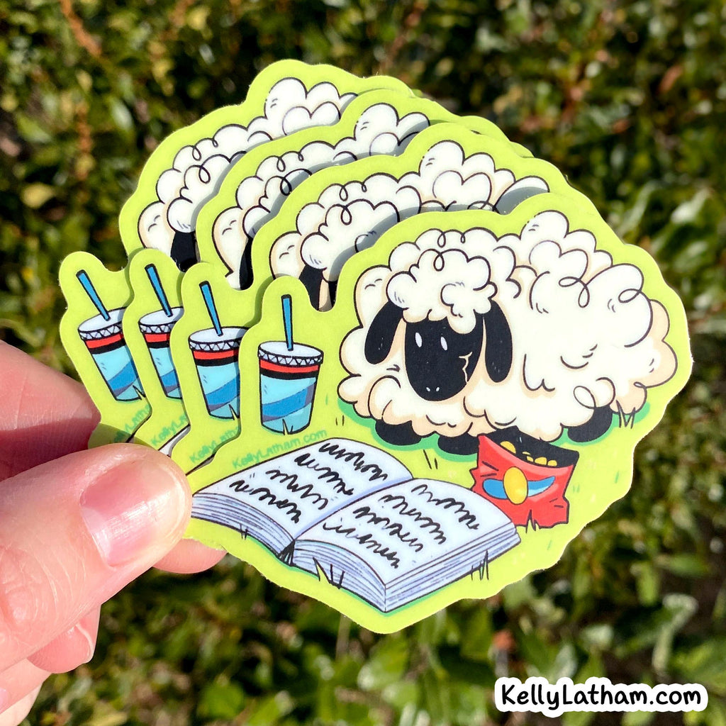 A hand holds a fan of die-cut stickers featuring a cartoon sheep, three striped drinks with straws, and an open book with handwritten text, against a blurred green outdoor background. A 'KellyLatham.