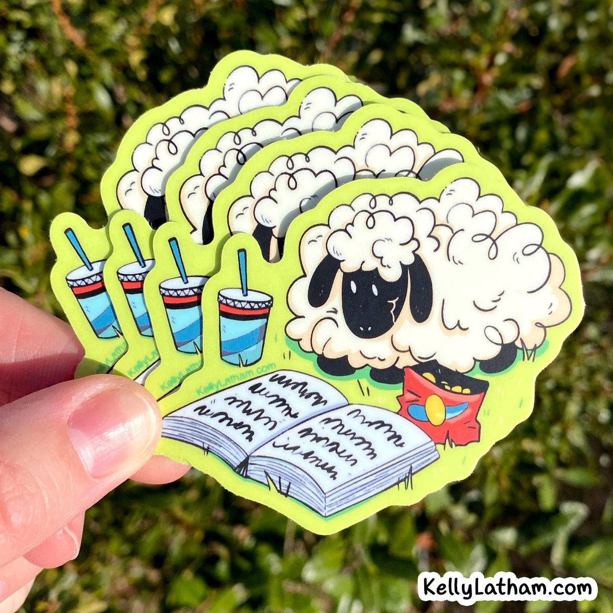 A hand holds a fan of die-cut stickers featuring a cartoon sheep, three striped drinks with straws, and an open book with handwritten text, against a blurred green outdoor background. A 'KellyLatham.