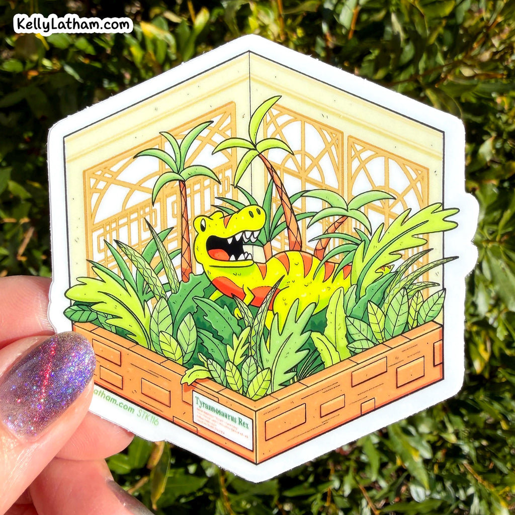 A hand with glittery purple nail polish holds a hexagonal die-cut sticker featuring a cartoon green dinosaur in a tropical jungle scene. The sticker is photographed outdoors against a background of green foliage.