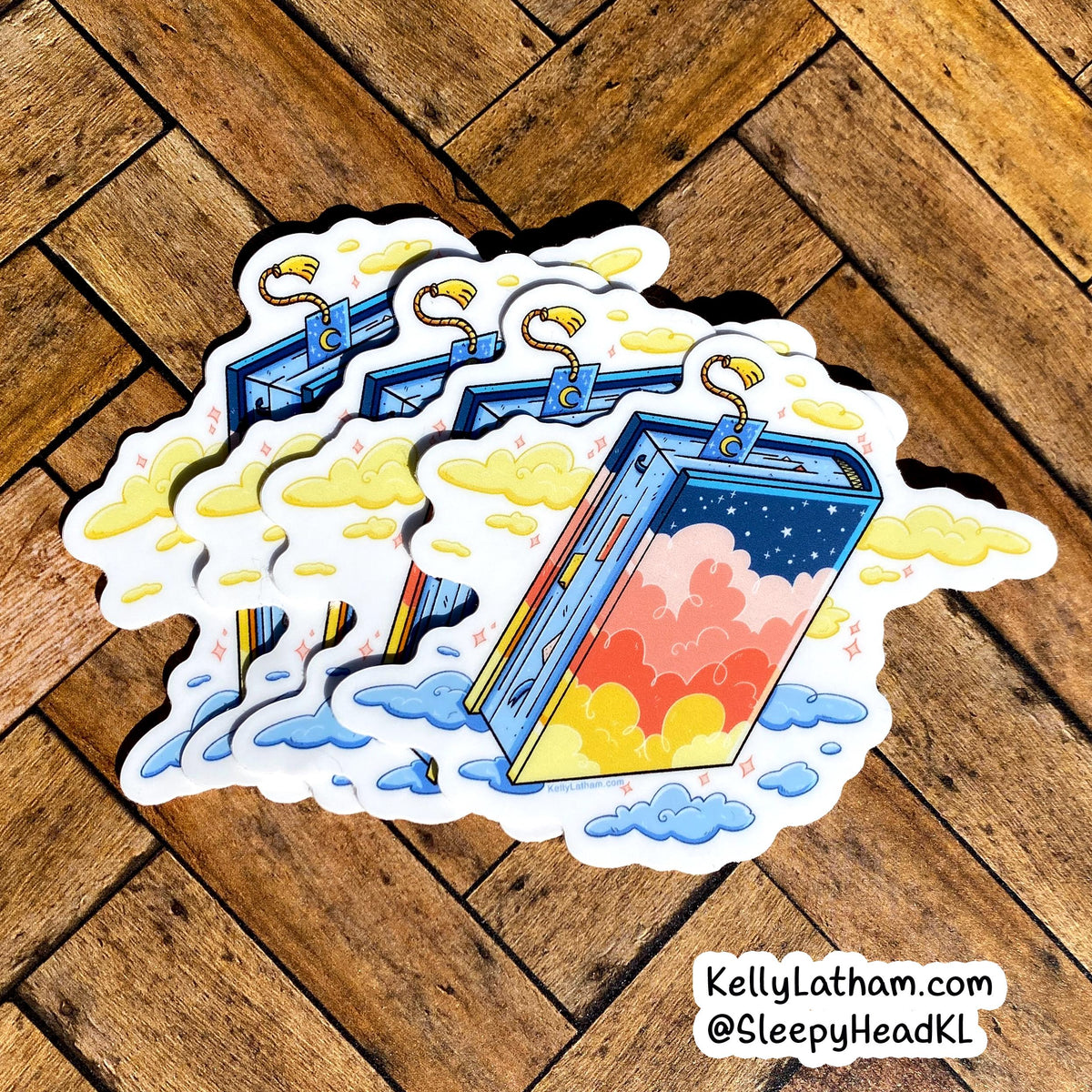 Three cartoon phone-shaped stickers with colorful sunset gradients, clouds, and rainbow accents, arranged on a wooden floor. A small watermark in the bottom right reads KellyLatham.