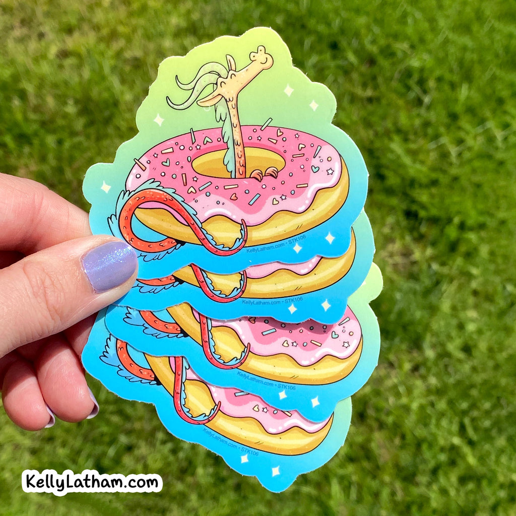 A hand with lavender nail polish holds a stack of die-cut stickers featuring a whimsical cartoon illustration of a long neck monster rising from pink-frosted donuts with sprinkles, set against a blurred green grass background. A watermark in the bottom-left corner reads 'KellyLatham.