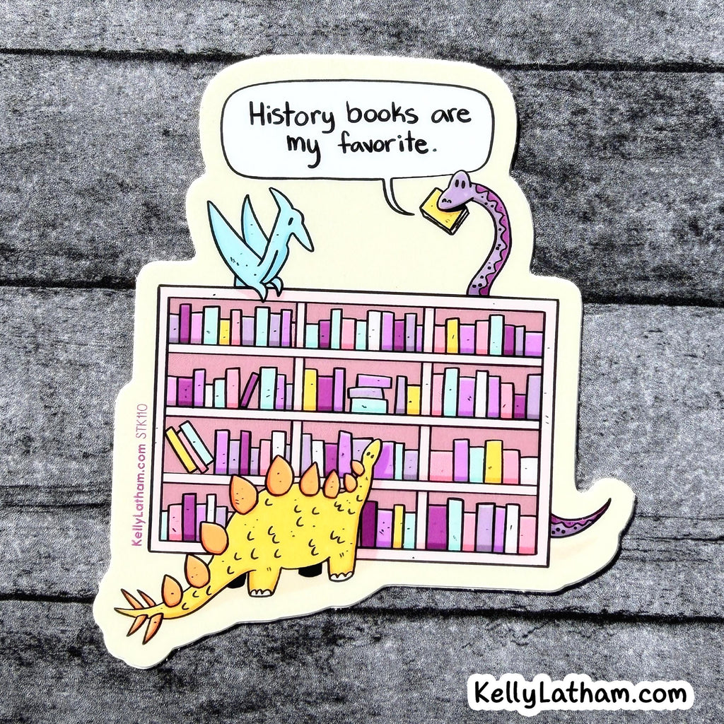 A die-cut sticker featuring a bookshelf with colorful books and three cartoon dinosaurs, with a speech bubble reading 'History books are my favorite.' The sticker is photographed on a gray wood-textured background.