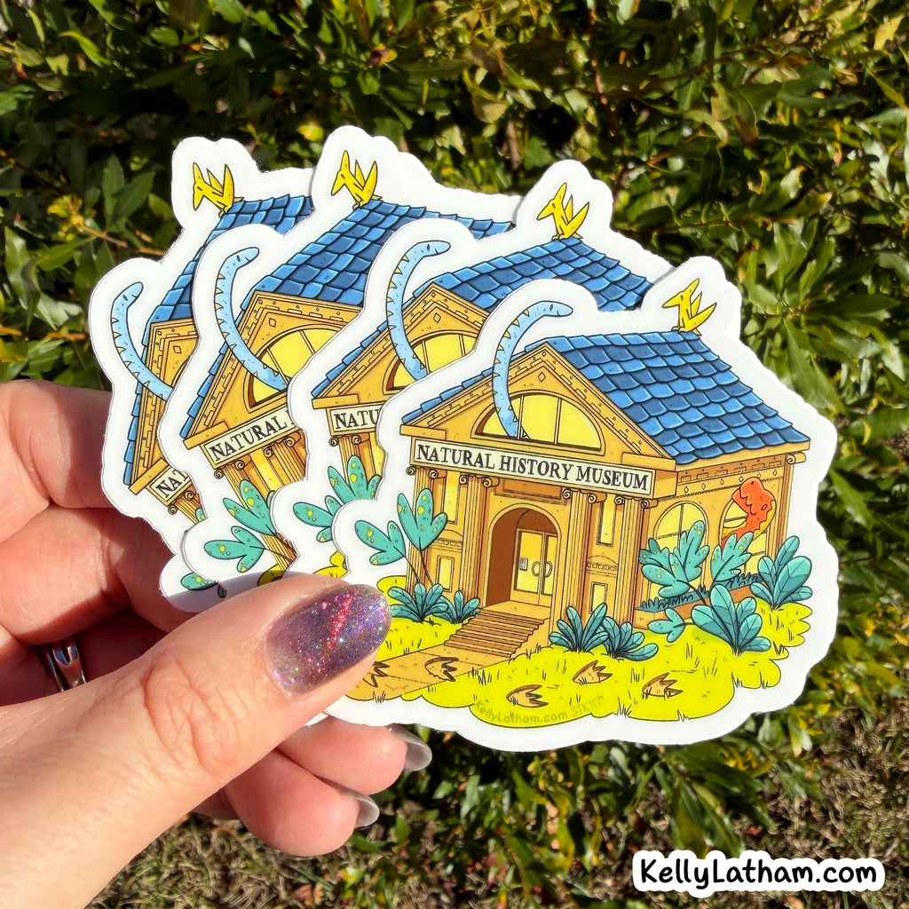 A hand holds three die-cut stickers featuring an illustrated natural history museum building with a blue roof, yellow walls, and green plants, against a background of green foliage.