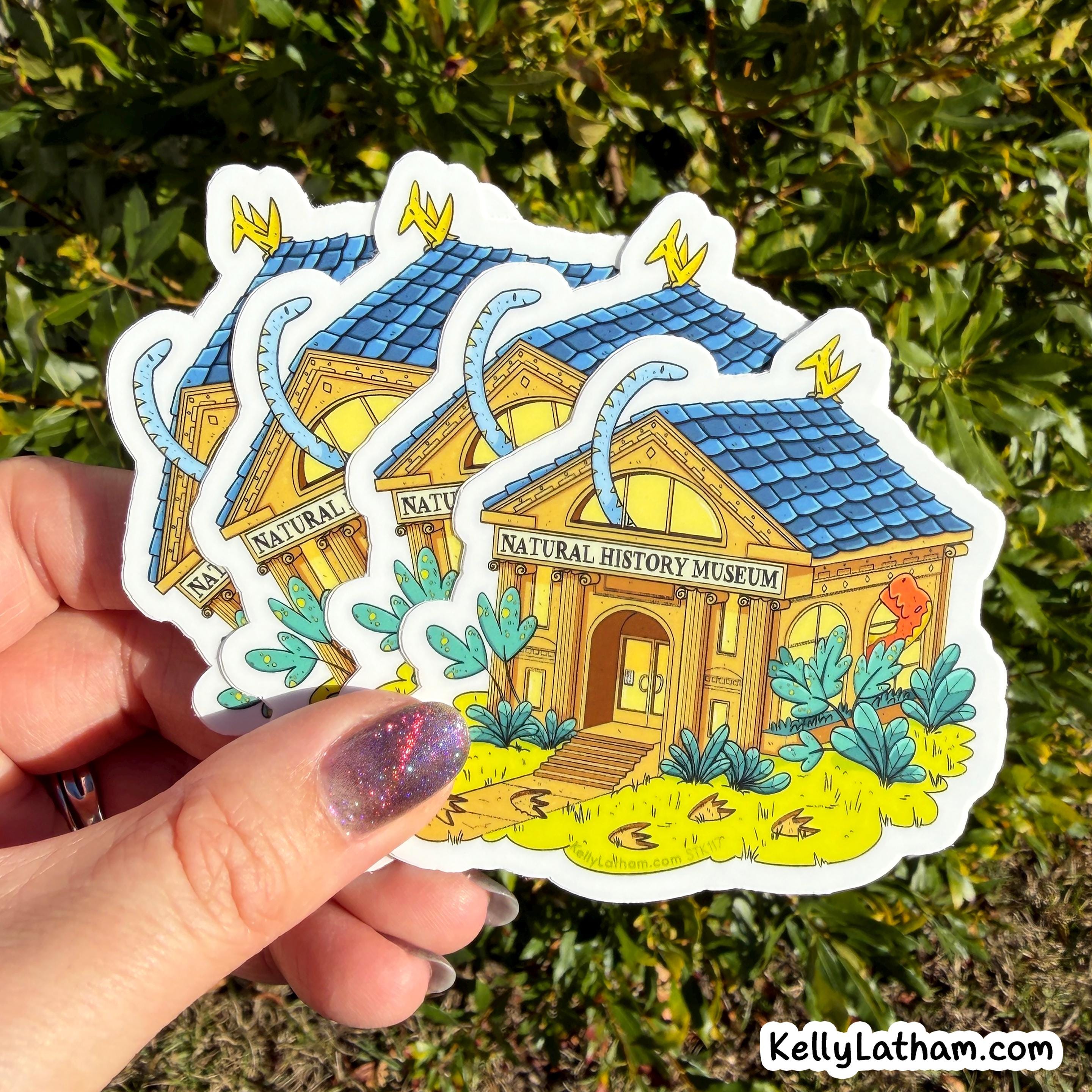 A hand holds three die-cut stickers featuring an illustrated natural history museum building with a blue roof, yellow walls, and green plants, against a background of green foliage.