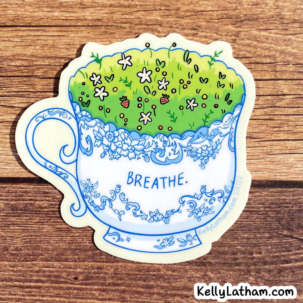 A die-cut sticker featuring a white teacup with blue floral detailing, filled with green grass and small wildflowers, placed on a brown wood background. The word 'BREATHE.