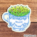 A die-cut sticker featuring a white teacup with blue floral detailing, filled with green grass and small wildflowers, placed on a brown wood background. The word 'BREATHE.