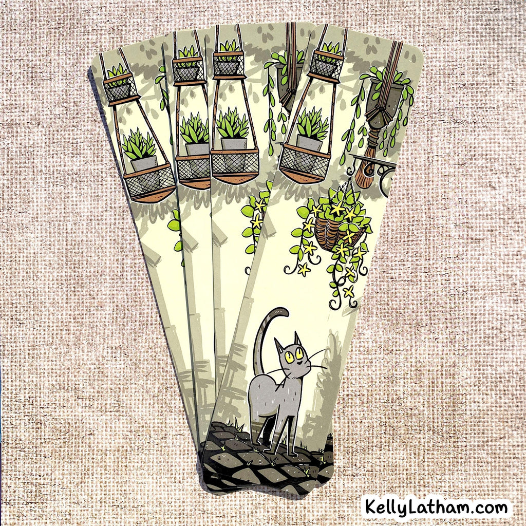 A decorative fan-shaped card or art print featuring an illustrated gray cat standing on a dark surface beneath hanging green plants and vines, set against a beige textured fabric background. The brand name KellyLatham.