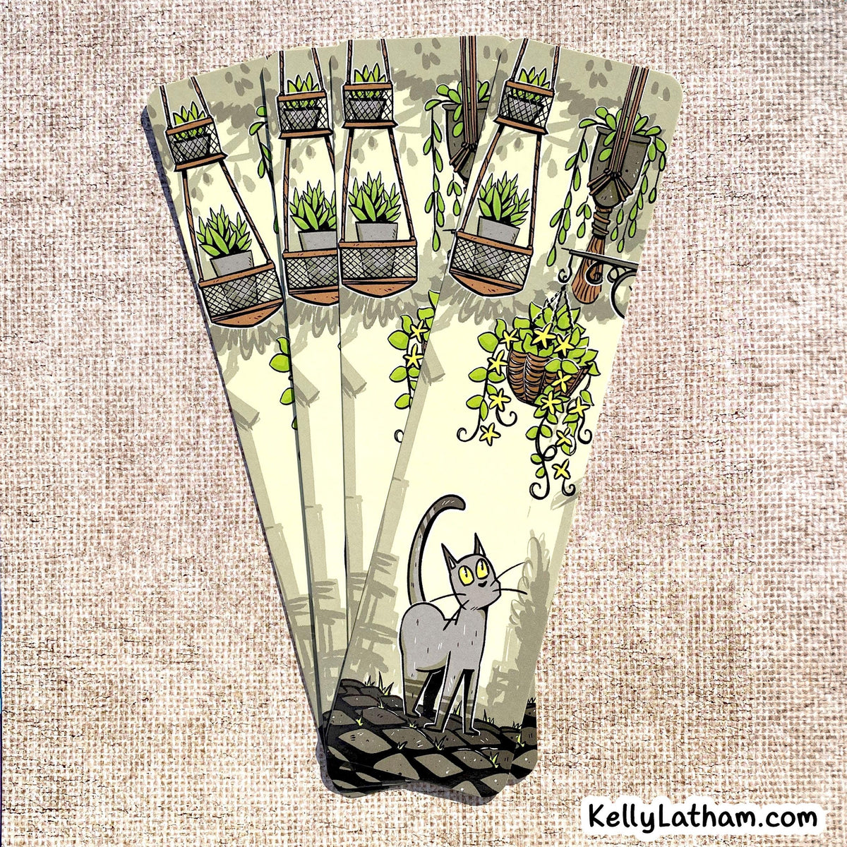 A decorative fan-shaped card or art print featuring an illustrated gray cat standing on a dark surface beneath hanging green plants and vines, set against a beige textured fabric background. The brand name KellyLatham.