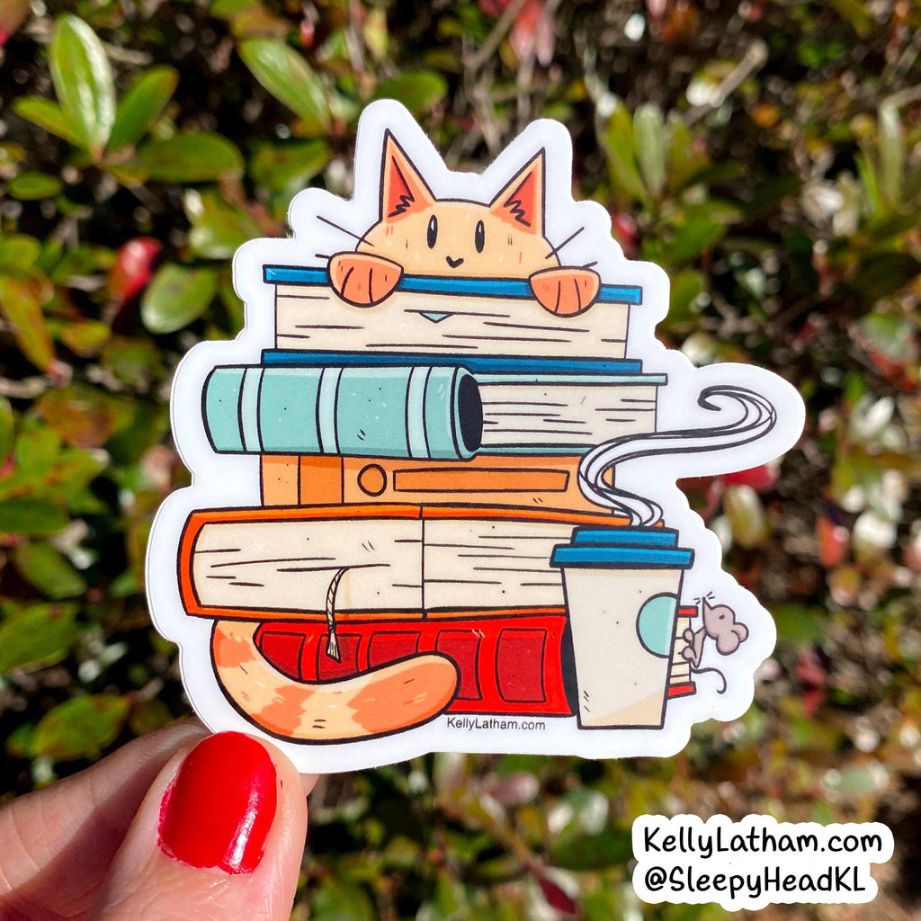 A die-cut sticker featuring a cute orange cat peeking over a stack of books, with a coffee cup and steam, held by a hand with red nail polish against a blurred green outdoor background. The sticker has the brand name KellyLatham printed on it.